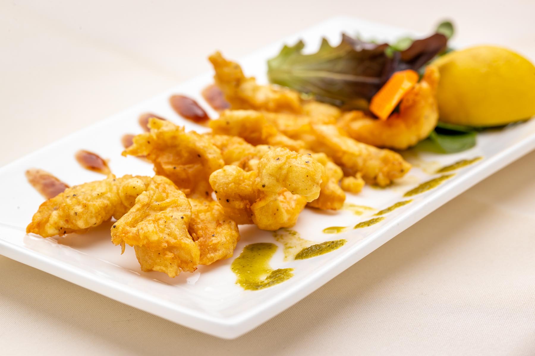 Golden fried battered shrimp on a white rectangular plate garnished with sauces, mixed greens, and a lemon wedge.