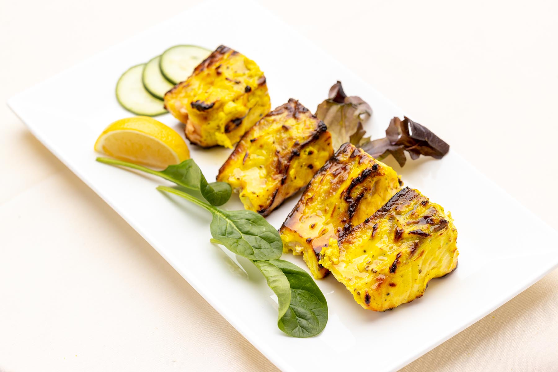 Grilled yellow marinated fish cubes served with lemon wedge, cucumber slices, and leafy greens on a white rectangular plate.