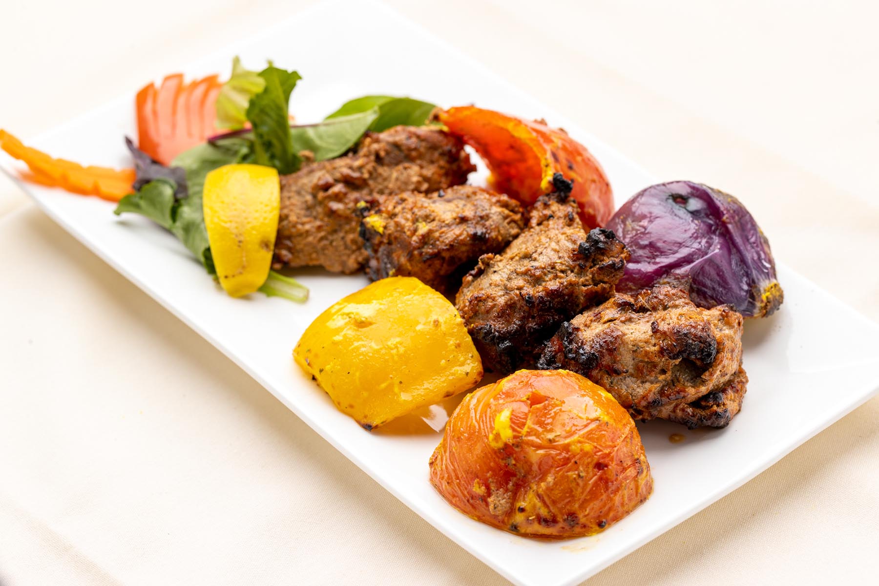 Grilled meat pieces served with roasted yellow bell pepper, tomato, red onion, and mixed greens on a white rectangular plate.