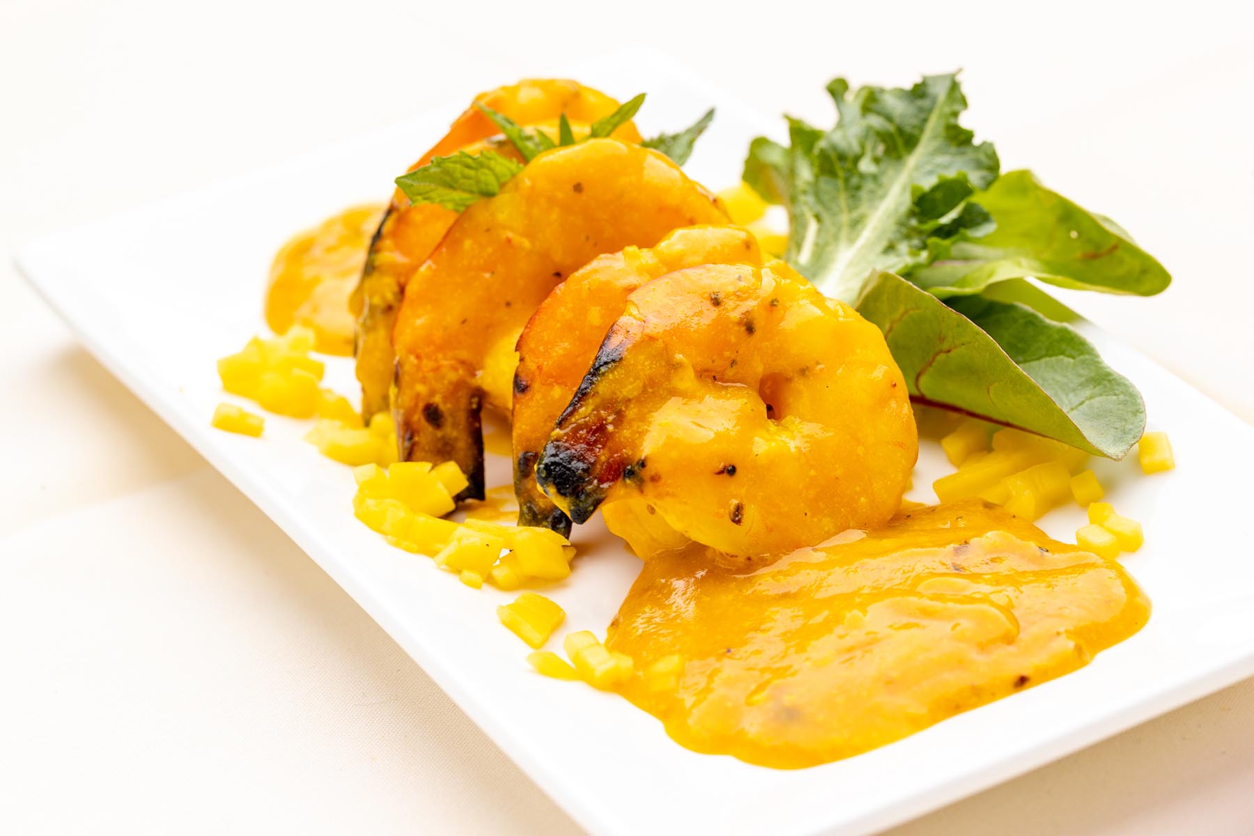 Grilled shrimp coated in a yellow sauce served on a white plate with chopped yellow garnish and green leafy salad.