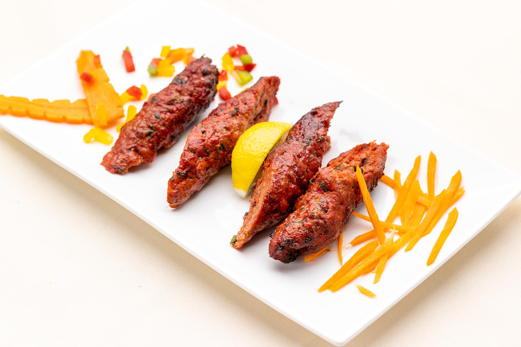 Four grilled seekh kebabs garnished with a lemon wedge, carrot sticks, and diced bell peppers on a white rectangular plate.