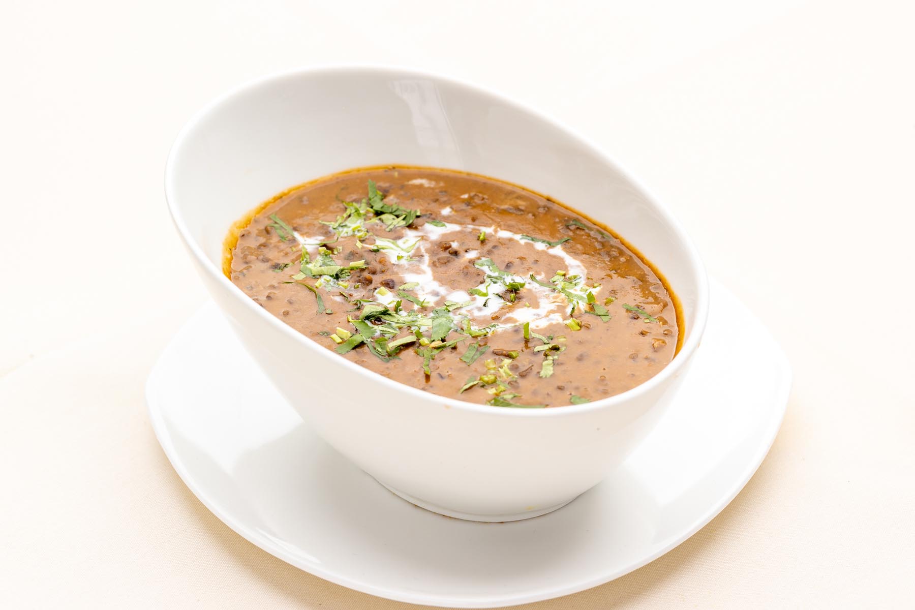 White bowl of dal makhani garnished with cream and chopped cilantro on a white plate.
