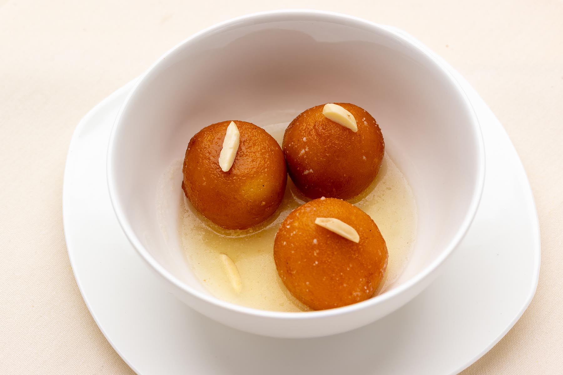 Three gulab jamuns garnished with almond slices in a white bowl with syrup.
