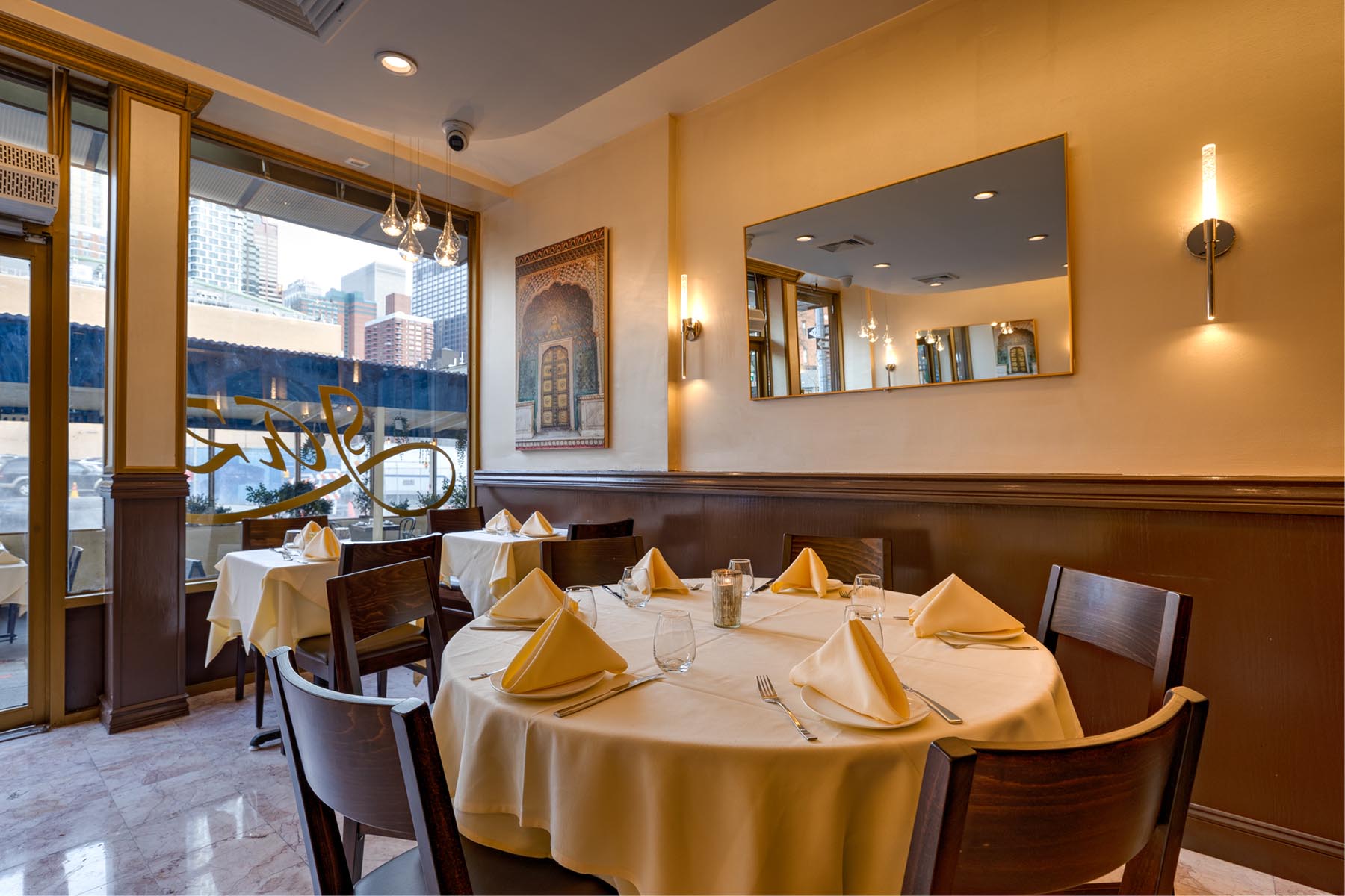 Cozy restaurant interior with a round table set with folded napkins, glassware, and cutlery, next to large windows and a wall mirror.