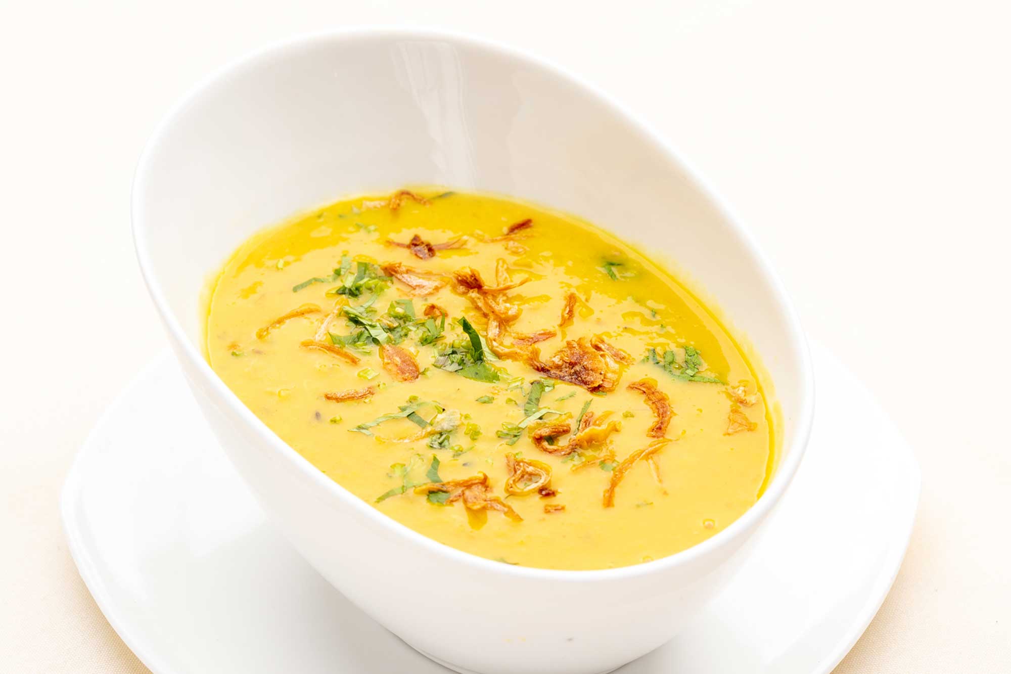 White bowl of yellow lentil soup garnished with fried onions and chopped cilantro on a white plate.
