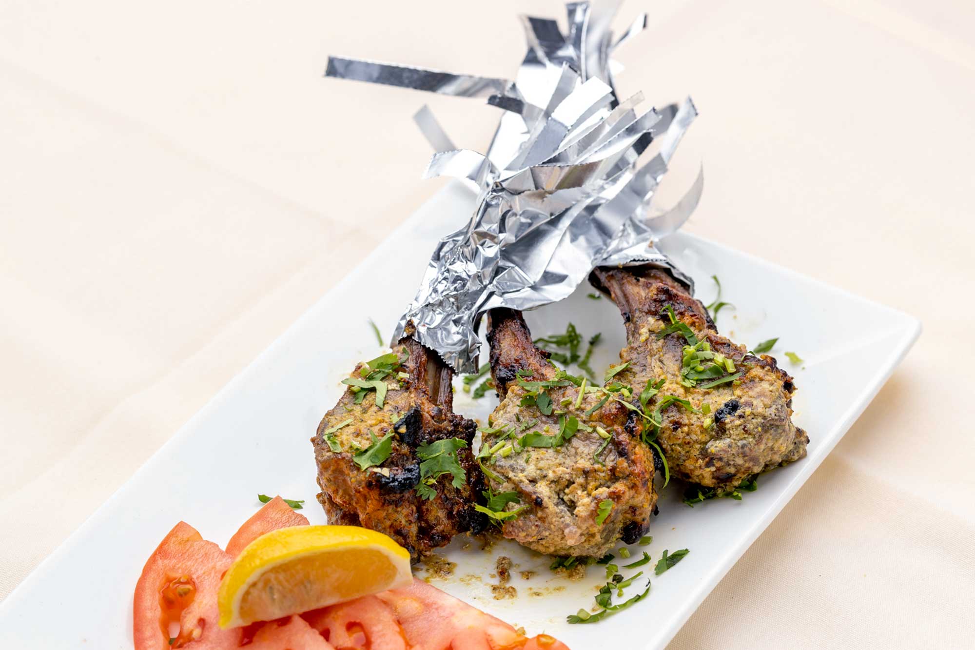 Three grilled lamb chops garnished with herbs and wrapped in foil at the bone ends, served with sliced tomatoes and a lemon wedge on a white plate.
