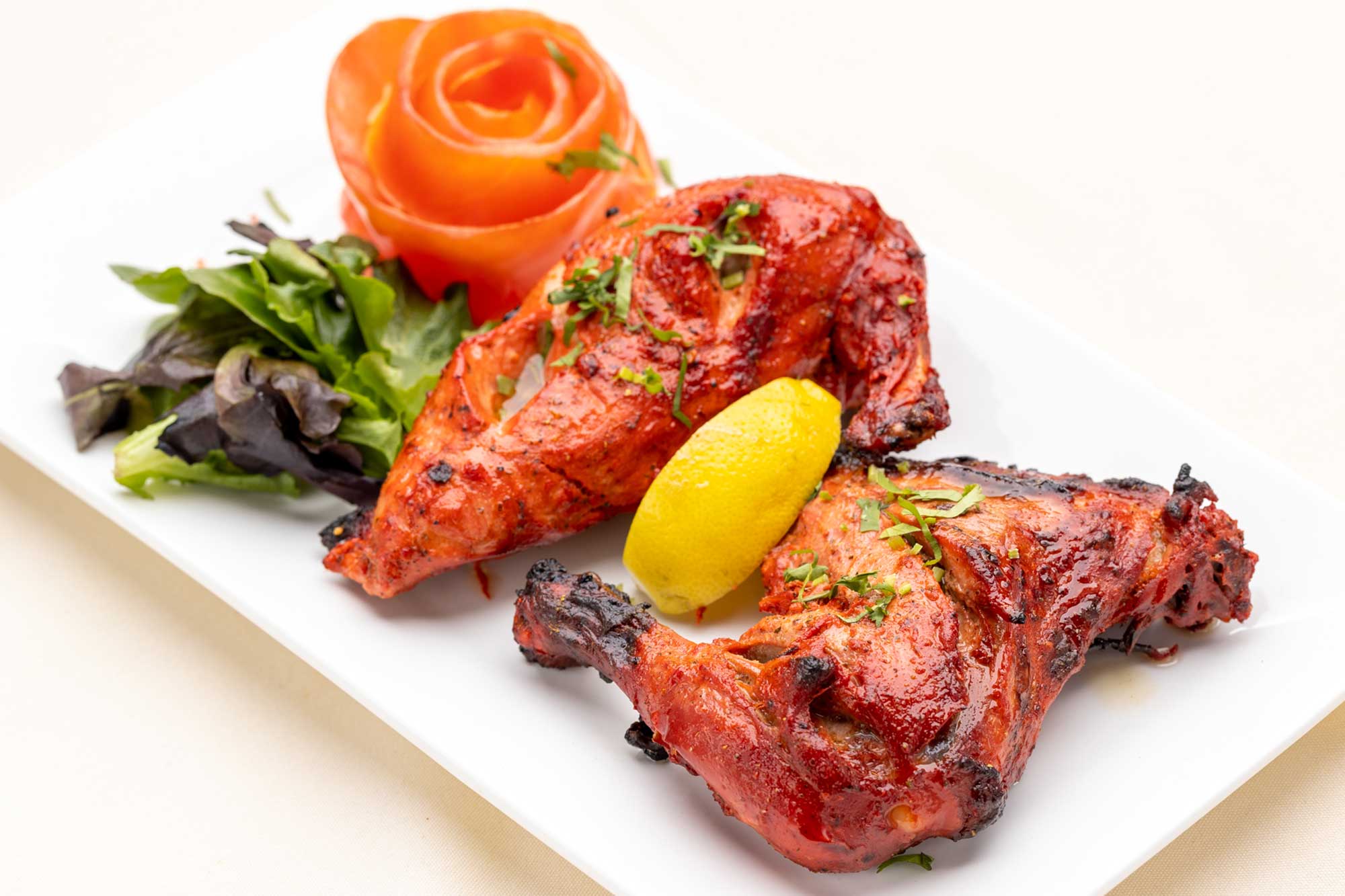 Two pieces of grilled tandoori chicken garnished with chopped herbs, served with a lemon wedge, lettuce, and a tomato rose on a white plate.