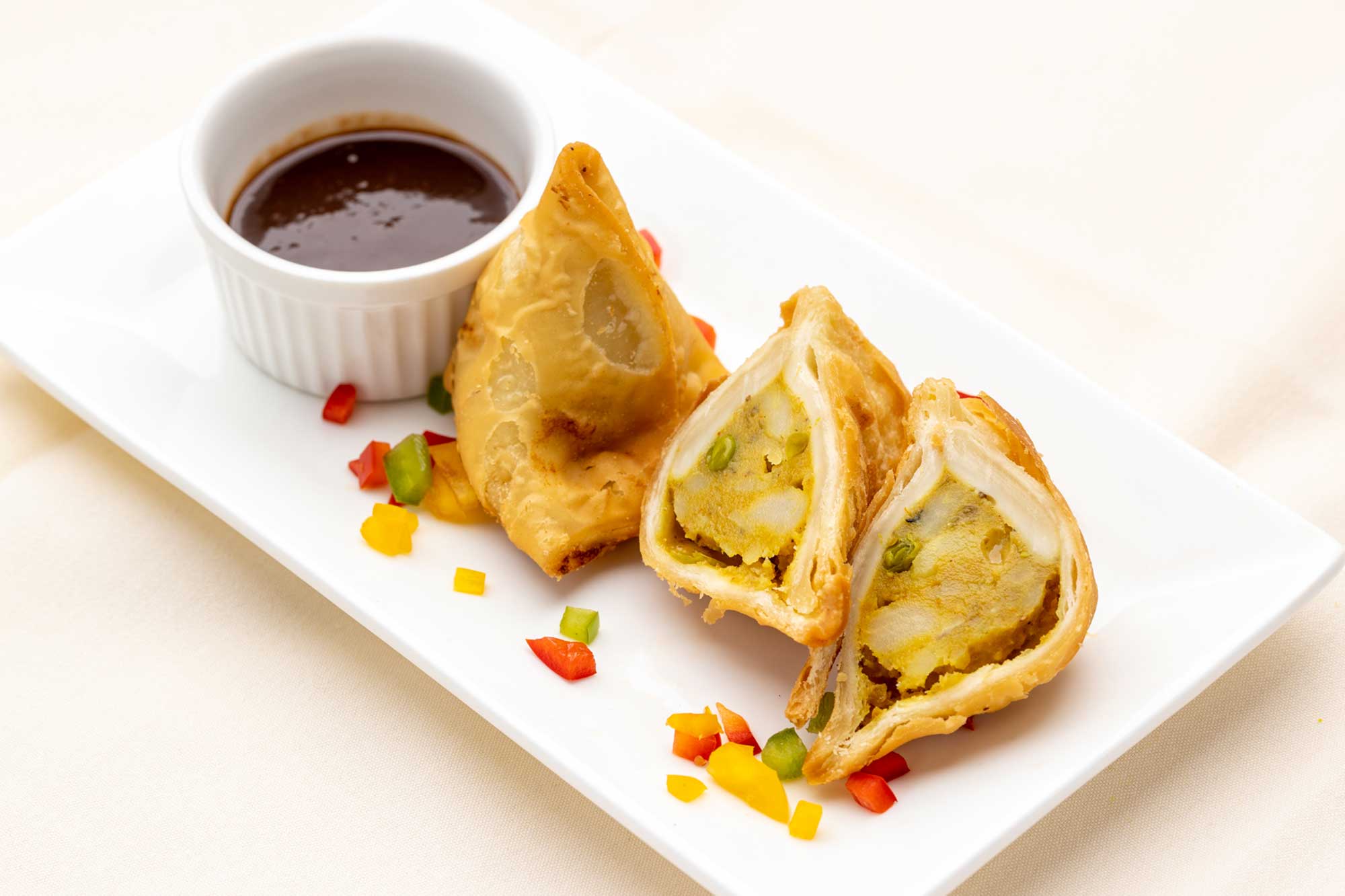 Golden-brown samosas on a white rectangular plate with chopped red, yellow, and green bell peppers and a small bowl of dark dipping sauce.