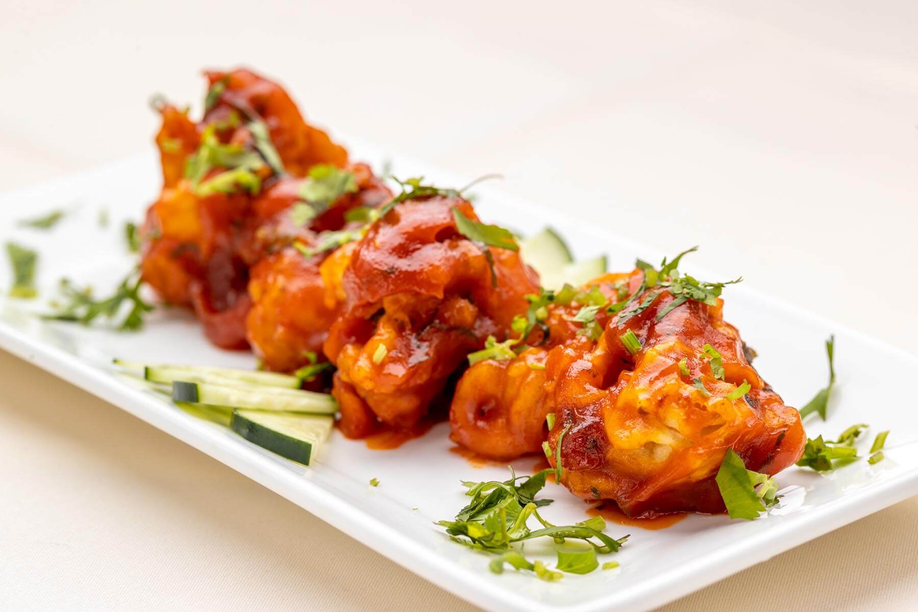 Four pieces of crispy fried chicken coated in red sauce and garnished with chopped herbs, served with cucumber slices on a white rectangular plate.