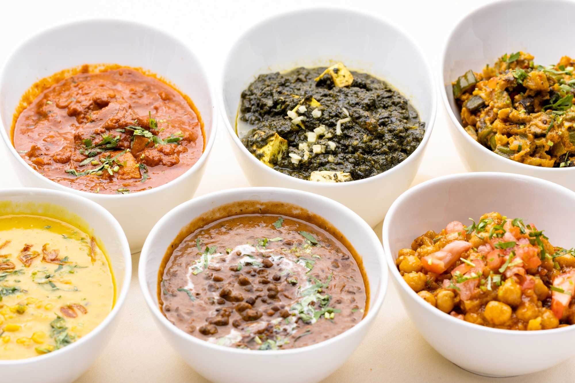 Six white bowls filled with assorted Indian dishes including dal, saag paneer, chole, and lentil curry garnished with herbs.