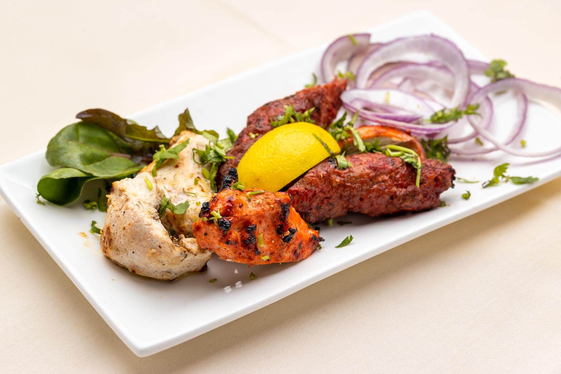 Plate with assorted Indian grilled meats, garnished with lemon wedge, fresh herbs, lettuce leaves, and sliced red onions.