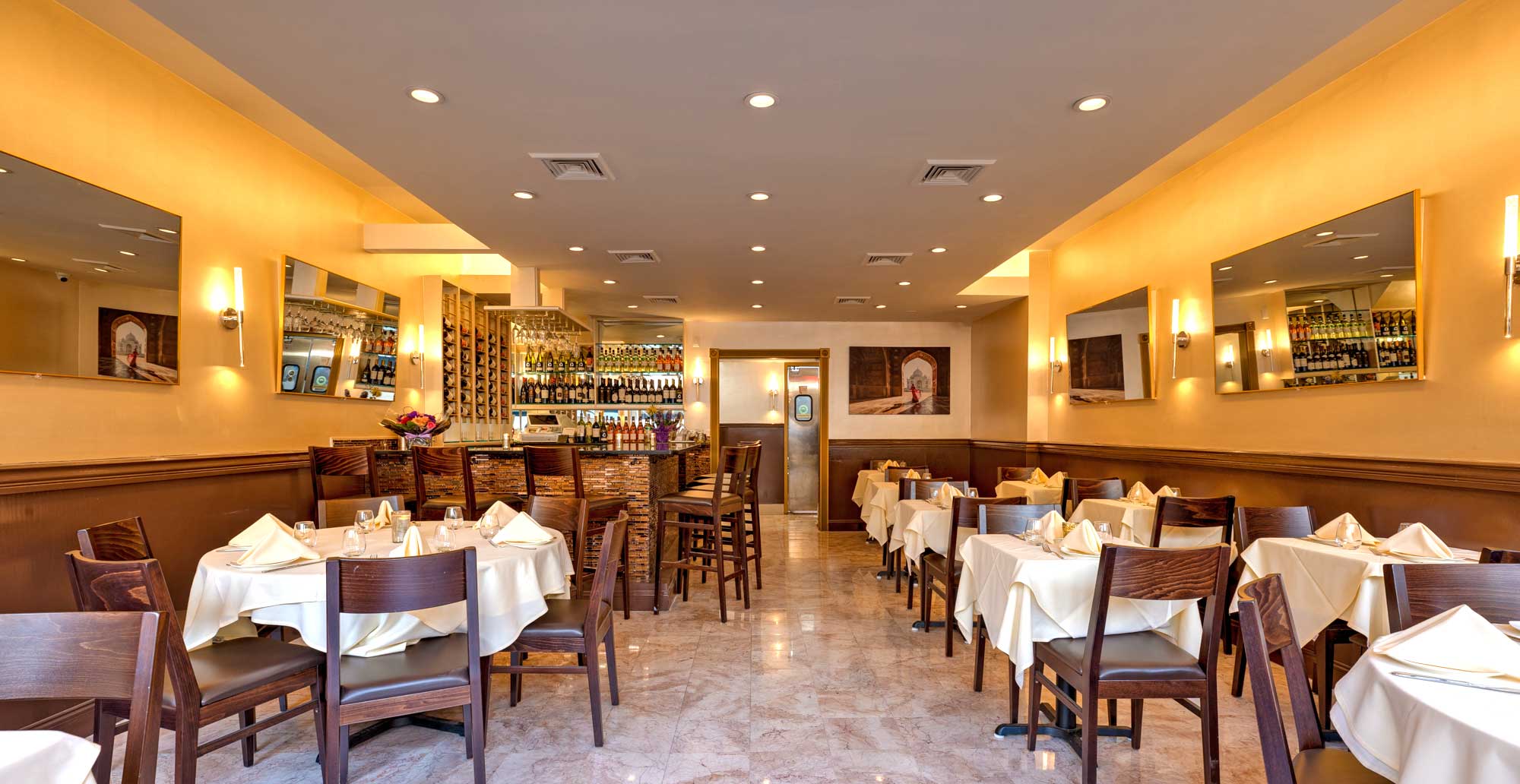 Elegant Indian restaurant interior with neatly set tables, wooden chairs, a bar with wine bottles, and warm lighting.
