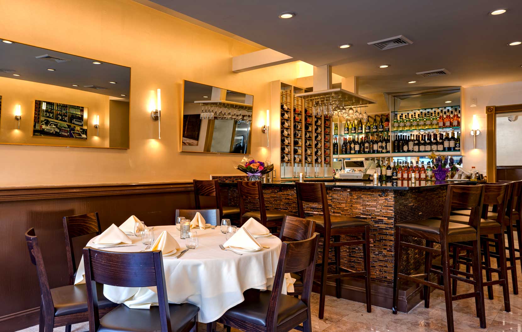 Cozy restaurant interior with a round table set with white napkins and glasses, and a bar stocked with wine bottles and glassware.