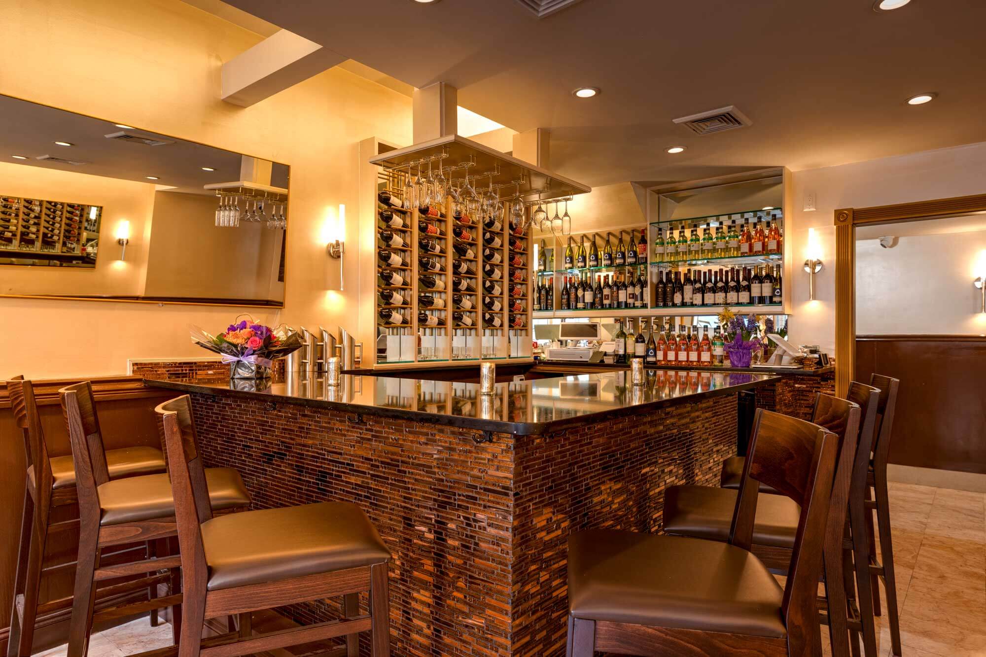 Modern bar with a dark polished countertop, wooden high chairs, wine bottles arranged on racks, and hanging wine glasses under warm lighting.
