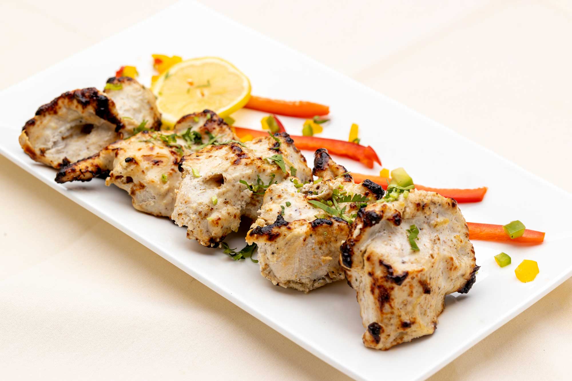 Grilled chicken pieces garnished with herbs served on a white plate with a lemon slice and sliced bell peppers.
