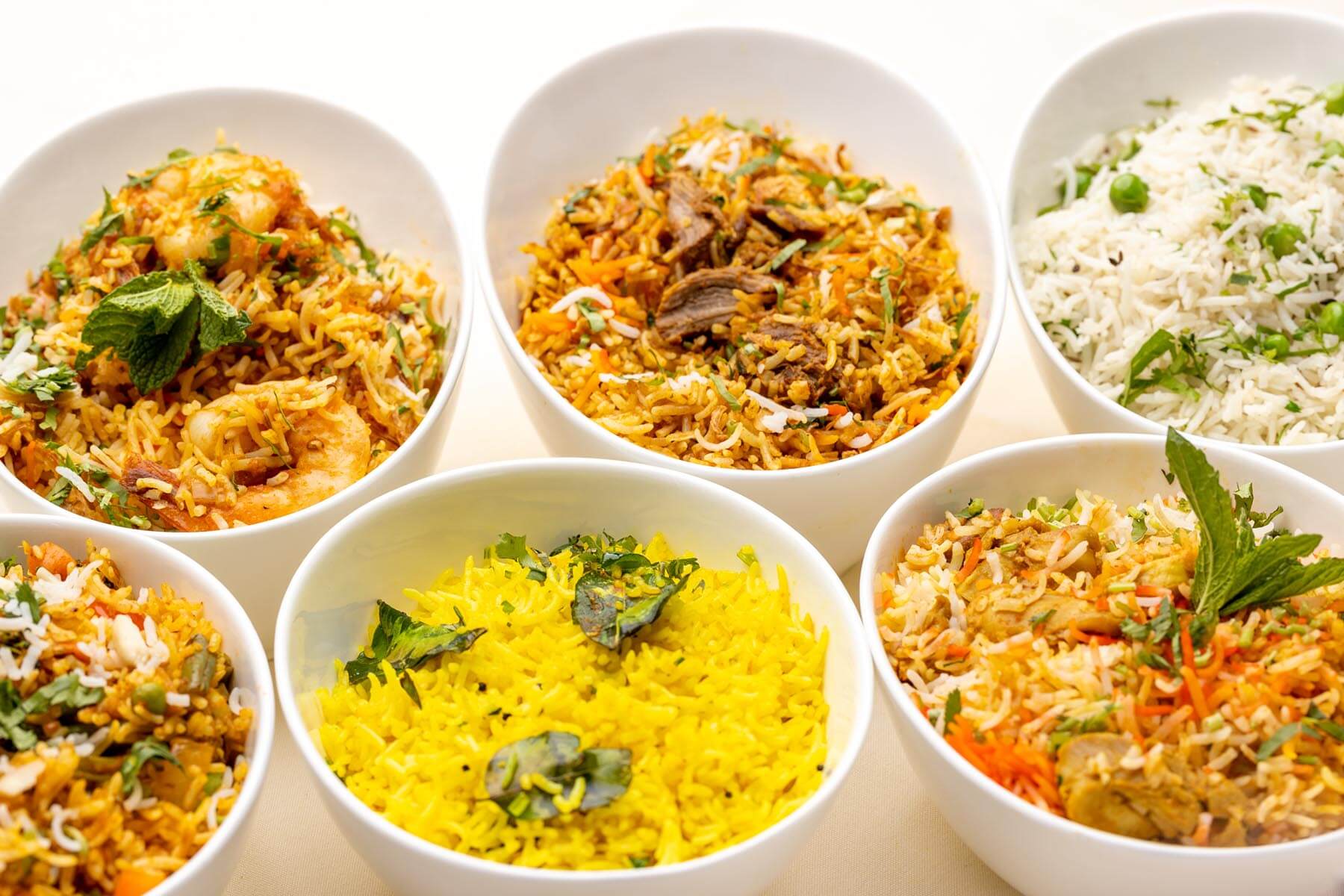 Six white bowls filled with various colorful Indian rice dishes garnished with herbs and spices.