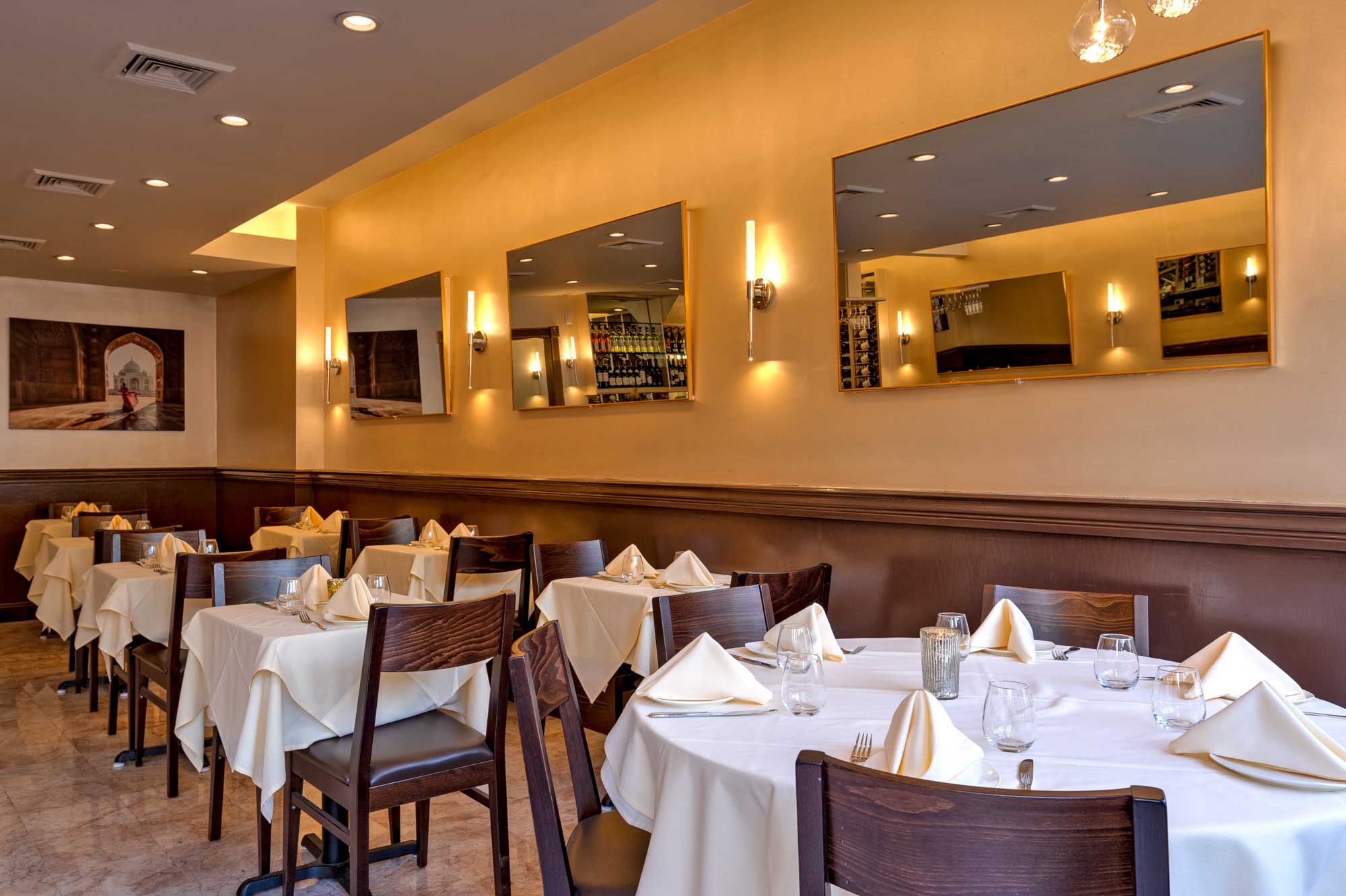 Elegant dining room with tables covered in white tablecloths, neatly folded napkins, wooden chairs, and wall mirrors reflecting a wine rack.