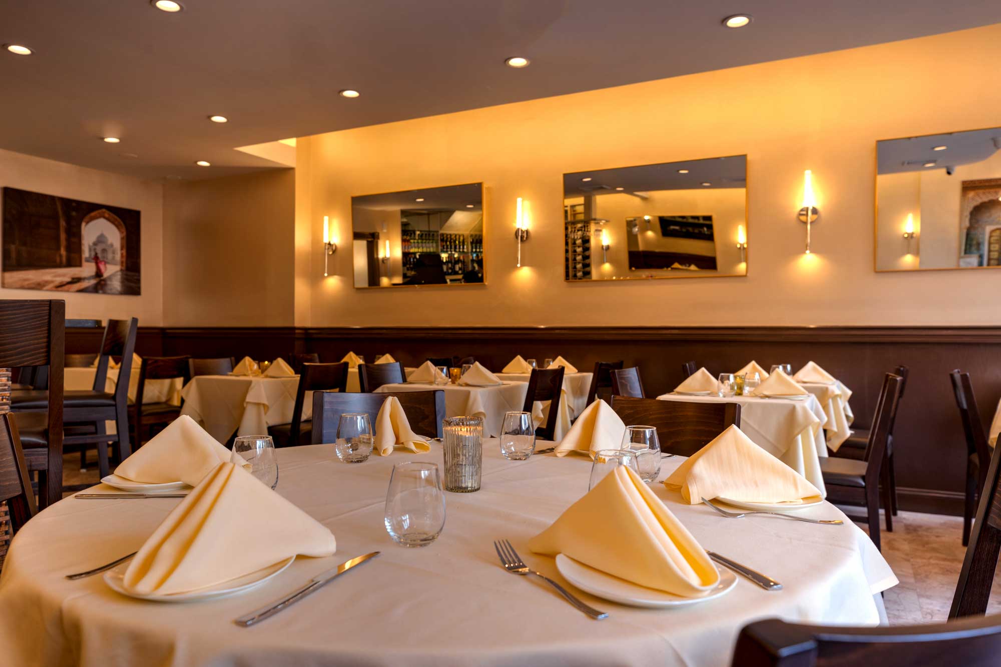 Elegant restaurant dining room with round tables set with beige napkins, glassware, and silverware under warm wall lighting.